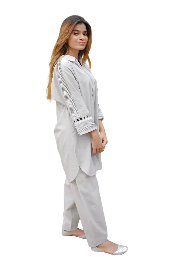 Woman in a light gray traditional outfit standing outdoors with greenery in the background
